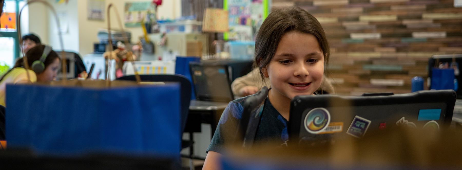 A young girl smiles while using a laptop in a bright classroom filled with colorful decorations.