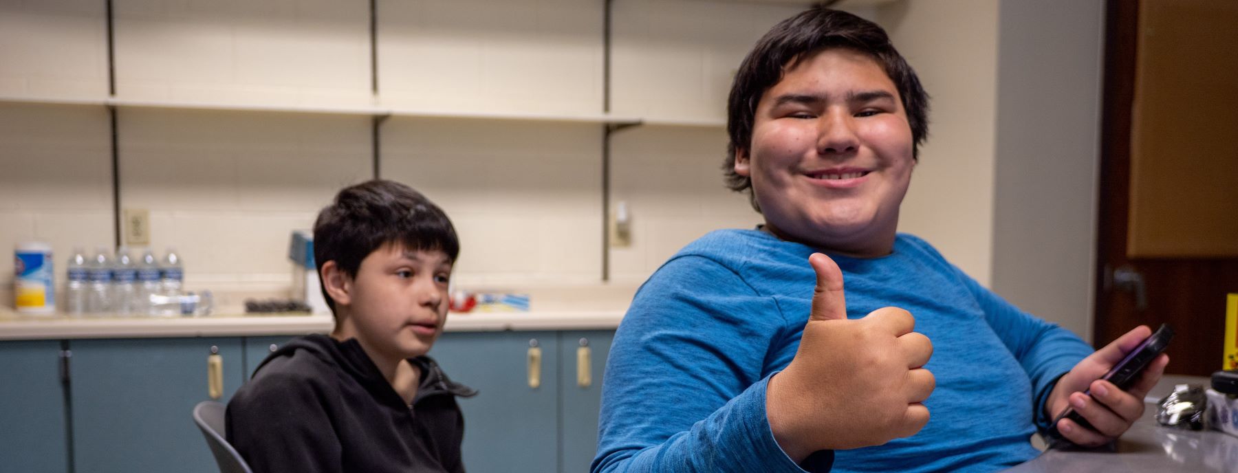 Two students at a table, one smiling and giving thumbs up to the camera.
